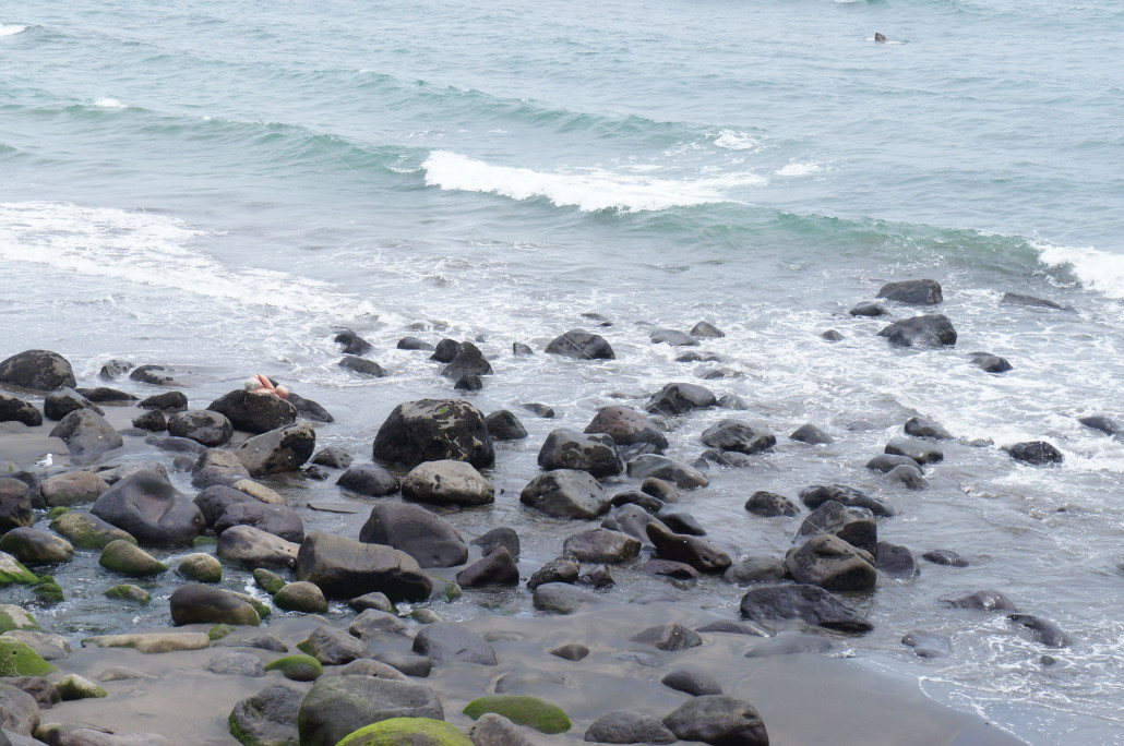 A beach with black boulders, and a distant figure lying on one of the boulders.