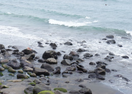 A beach with black boulders, and a distant figure lying on one of the boulders.