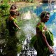 Women stand in a line in a beautiful blue lagoon waring leaves on their bodies and heads.