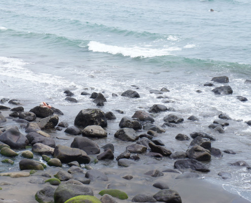 A beach with black boulders, and a distant figure lying on one of the boulders.