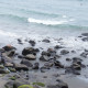 A beach with black boulders, and a distant figure lying on one of the boulders.