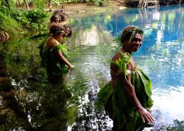 Women stand in a line in a beautiful blue lagoon waring leaves on their bodies and heads.