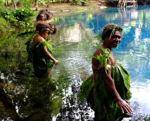 Women stand in a line in a beautiful blue lagoon waring leaves on their bodies and heads.