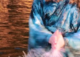 A still image of the motion of a woman in blue robes stands in water reflecting orange, and clapping her hand with the water between them.