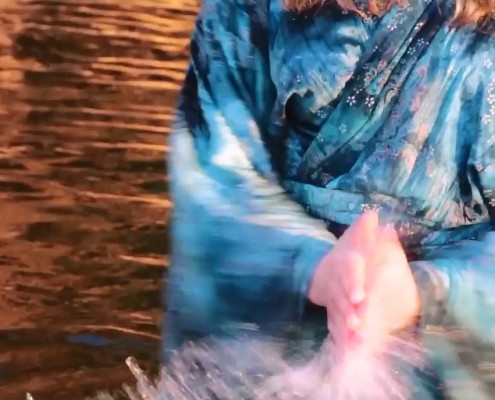 A still image of the motion of a woman in blue robes stands in water reflecting orange, and clapping her hand with the water between them.