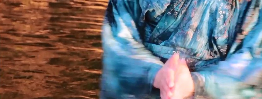 A still image of the motion of a woman in blue robes stands in water reflecting orange, and clapping her hand with the water between them.